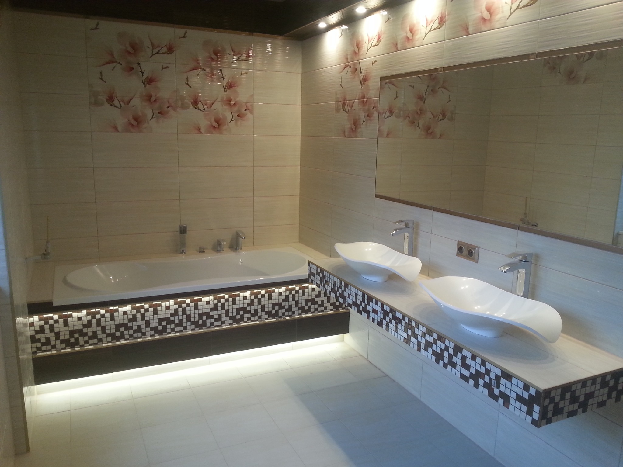 Elegancka łazienka z beżowymi płytkami zdobionymi motywem kwiatowym, podświetlaną wanną obudowaną mozaiką i umywalkami w kształcie łzy.