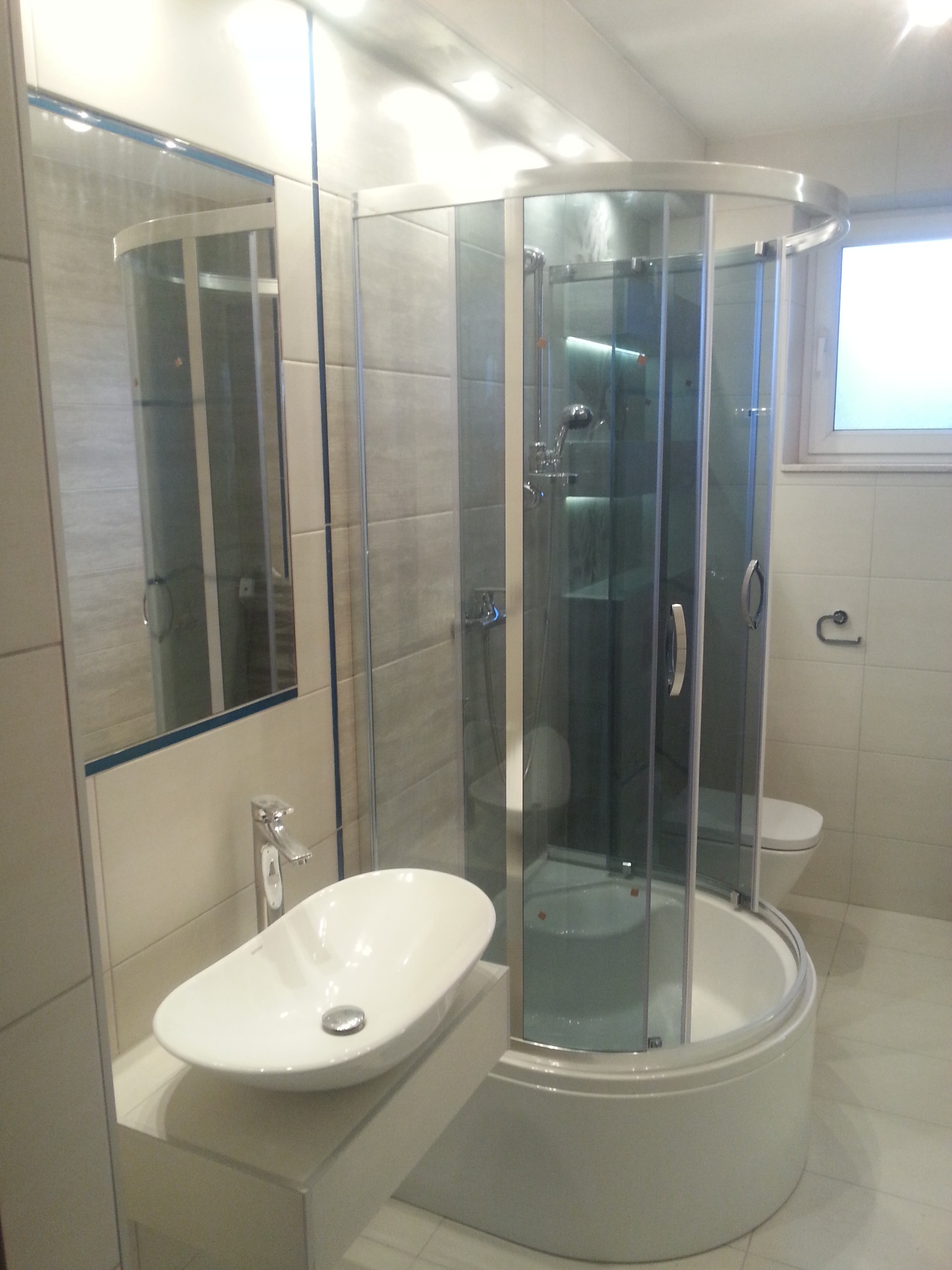 Nowoczesna łazienka z kabiną prysznicową z zaokrąglonymi szybami, umywalką nablatową i jasnymi płytkami.