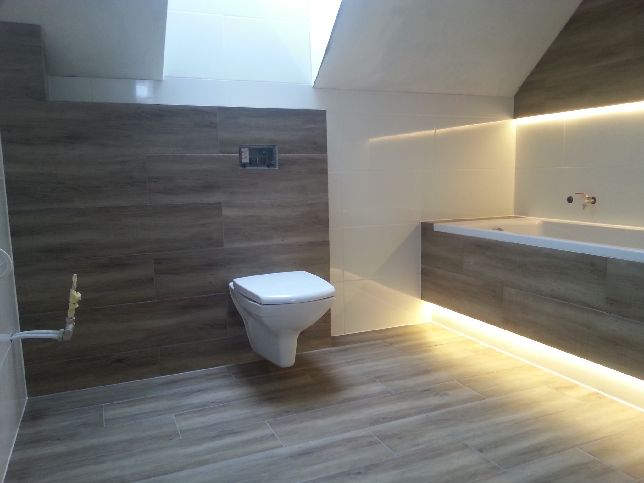 Wykończona łazienka na poddaszu z drewnopodobnymi płytkami na ścianach i podłodze, podwieszana toaleta, listwa LED pod wanną, oraz okno dachowe.