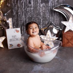 FotoŚliwka.pl - Uśmiechnięty roczny chłopiec w przezroczystej misce wypełnionej mleczną kąpielą, dekoracje urodzinowe w tle, sesja zdjęciowa w Chorzowie.