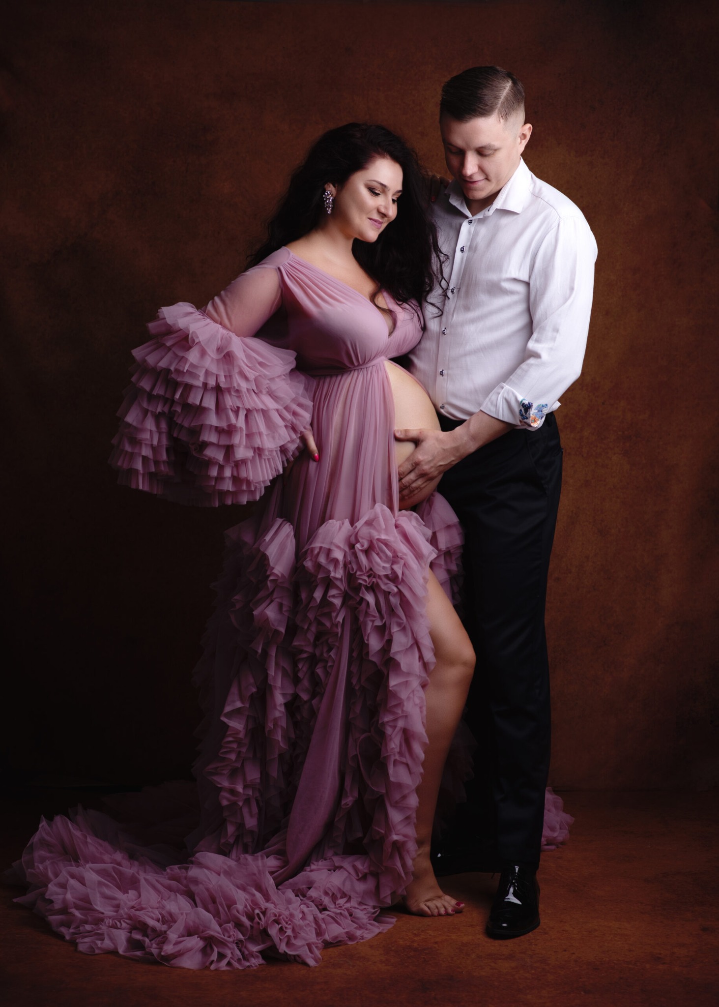 Kobieta w zwiewnej, falbaniastej sukni w kolorze pudrowego różu, z czarnymi kręconymi włosami i kolczykami, obejmowana przez mężczyznę w białej koszuli i czarnych spodniach, oboje patrzą na jej...