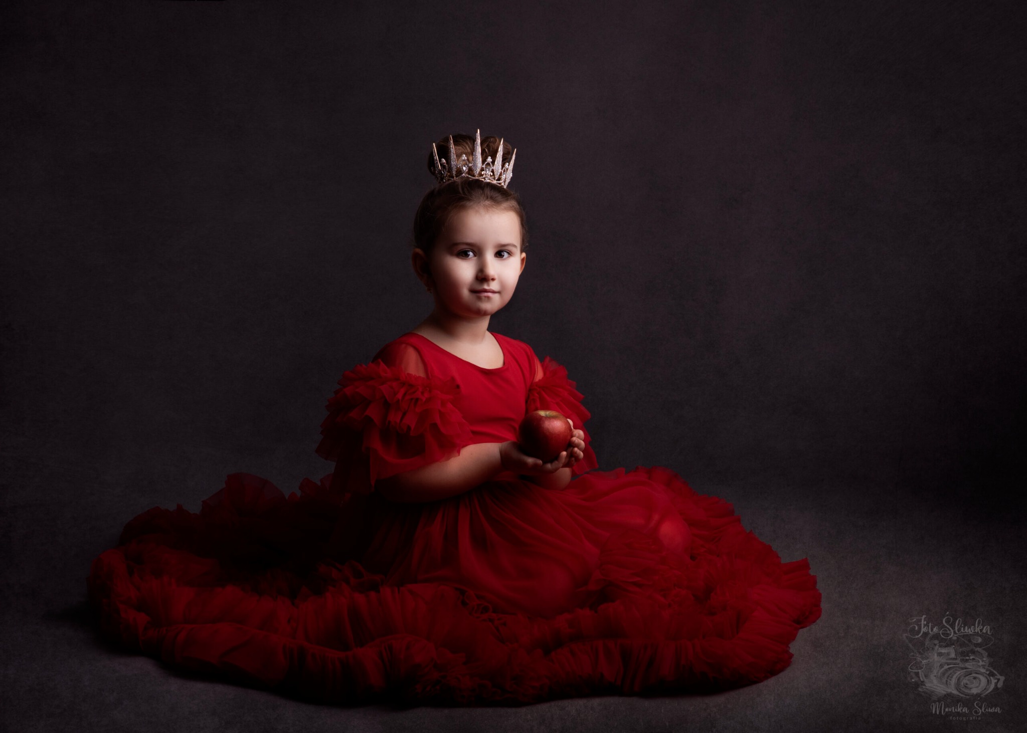 Dziewczynka w czerwonej, balowej sukni z falbanami i koroną na głowie trzyma czerwone jabłko na tle ciemnoszarego, lekko rozmytego tła.