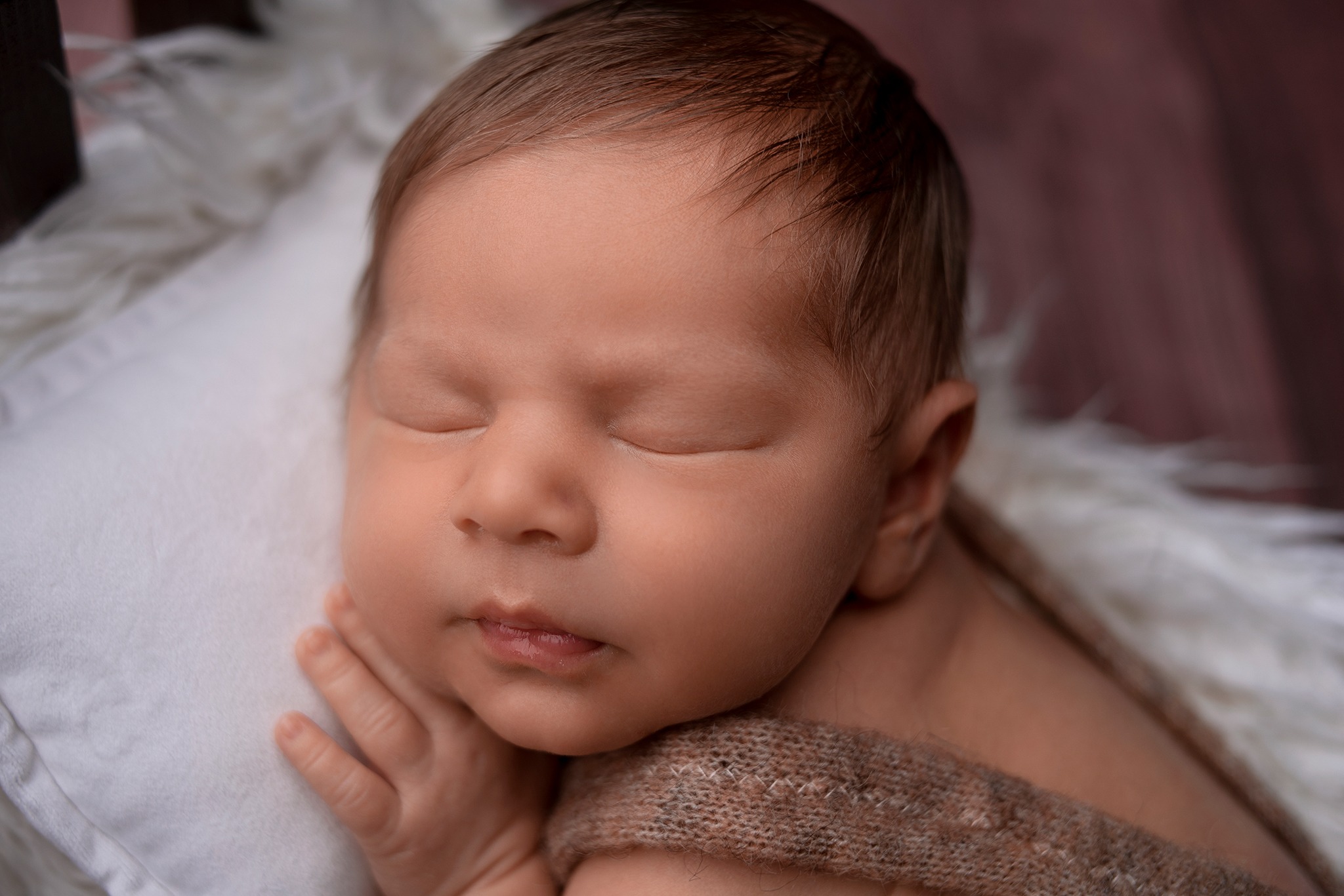 Śpiący noworodek z delikatnym meszkiem na głowie, otulony brązowym kocykiem na białym, puszystym podłożu