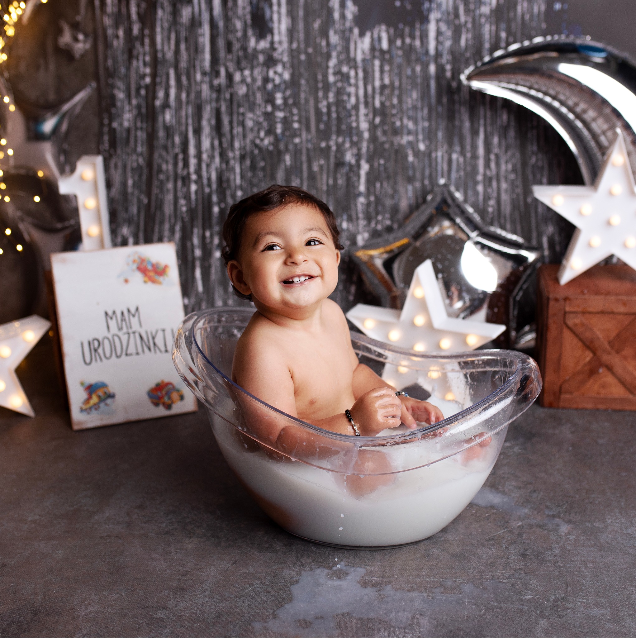 Uśmiechnięty roczny chłopiec w przezroczystej misce wypełnionej mleczną kąpielą, dekoracje urodzinowe w tle, sesja zdjęciowa w Chorzowie.