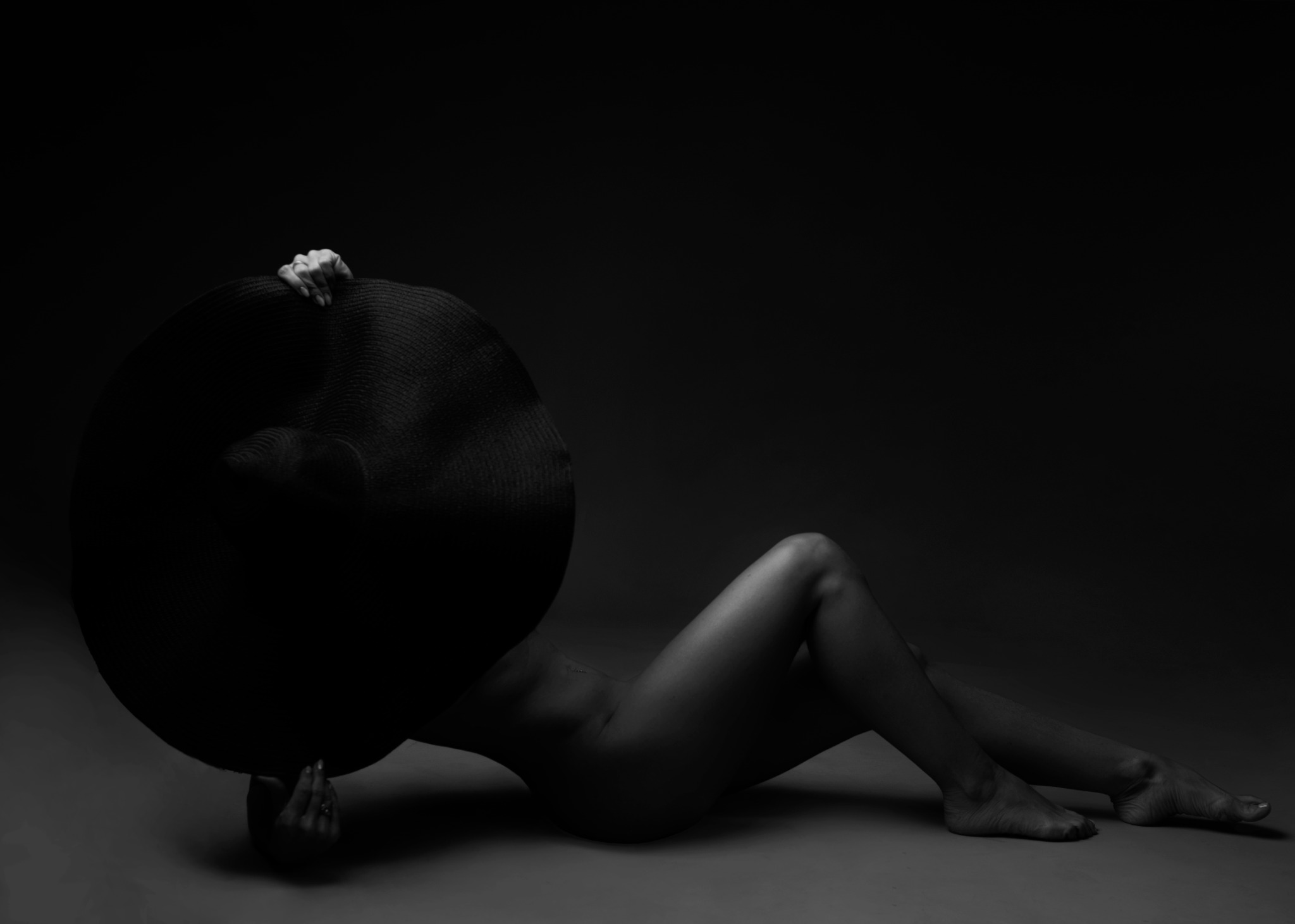 Czarno-białe zdjęcie kobiety leżącej na podłodze, zakrywającej twarz dużym, czarnym kapeluszem o szerokim rondzie; dłonie widoczne trzymają kapelusz, na palcu pierścionek.