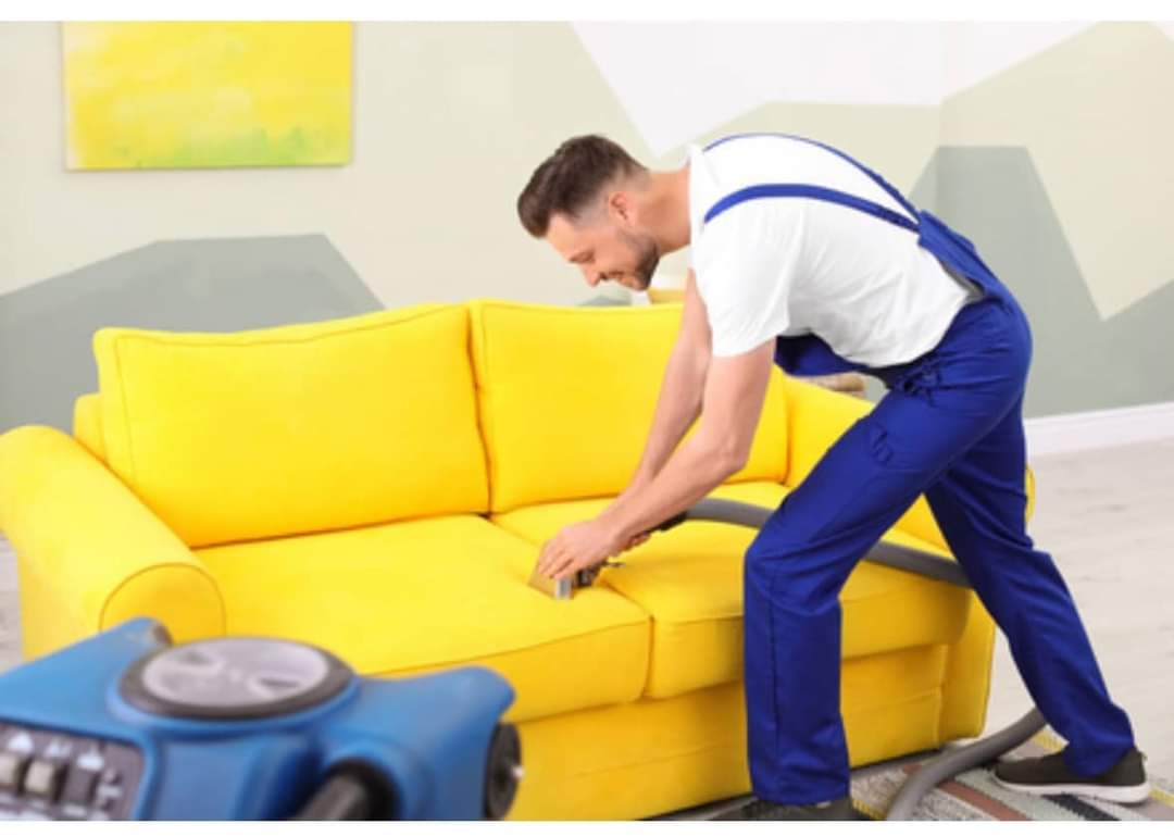 Mężczyzna w niebieskim kombinezonie czyści żółtą kanapę za pomocą niebieskiego odkurzacza piorącego. W tle widoczny obraz z abstrakcyjnym motywem.