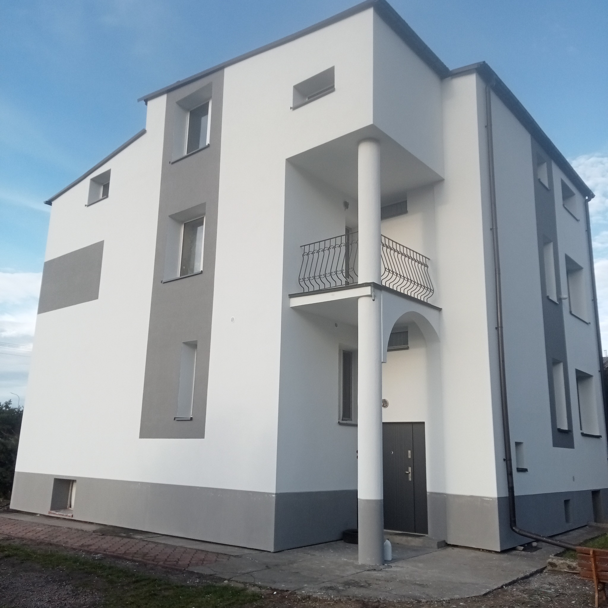 Nowoczesny dom po malowaniu elewacji w odcieniach bieli i szarości, z widocznym balkonem z ozdobną balustradą i kolumną.