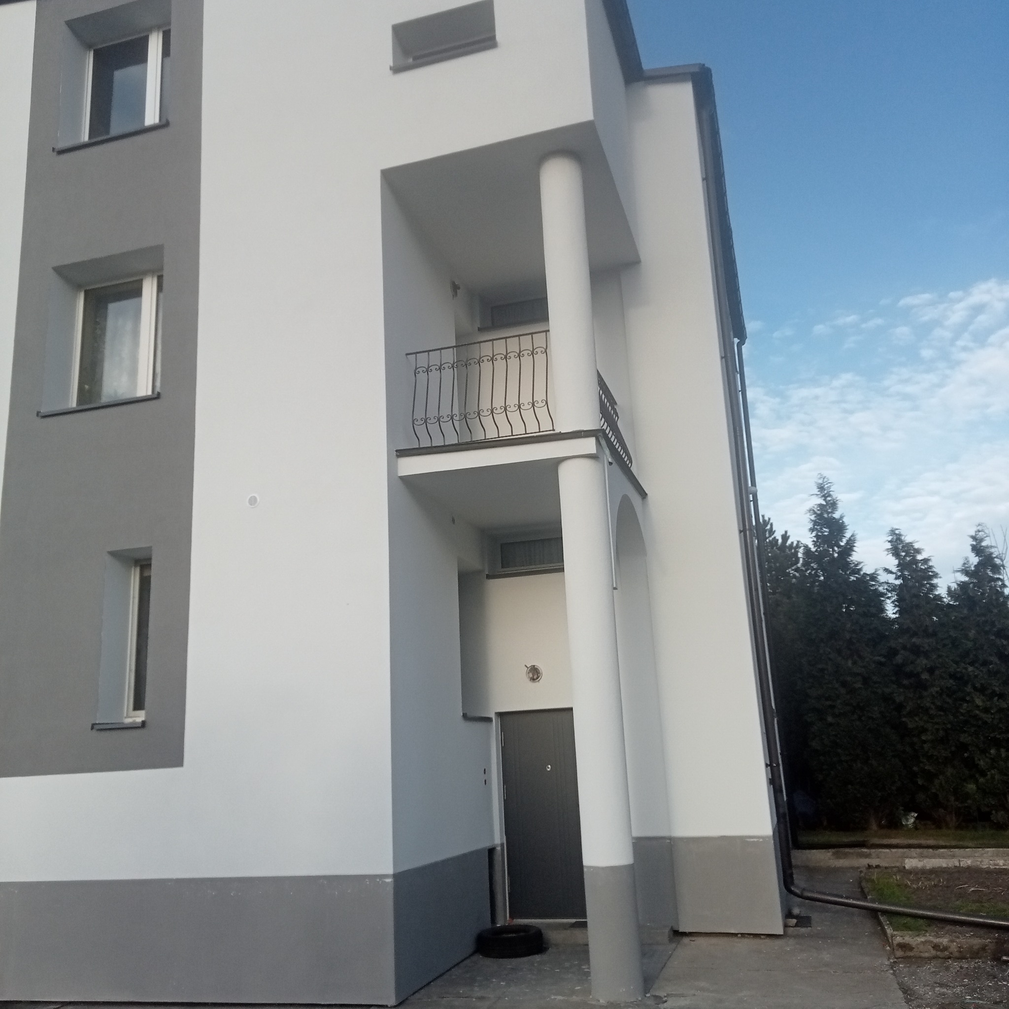 Nowoczesny budynek po zakończonych pracach elewacyjnych, widoczne biało-szare tynki, balkon z ozdobną balustradą i ciemne drzwi wejściowe na parterze.