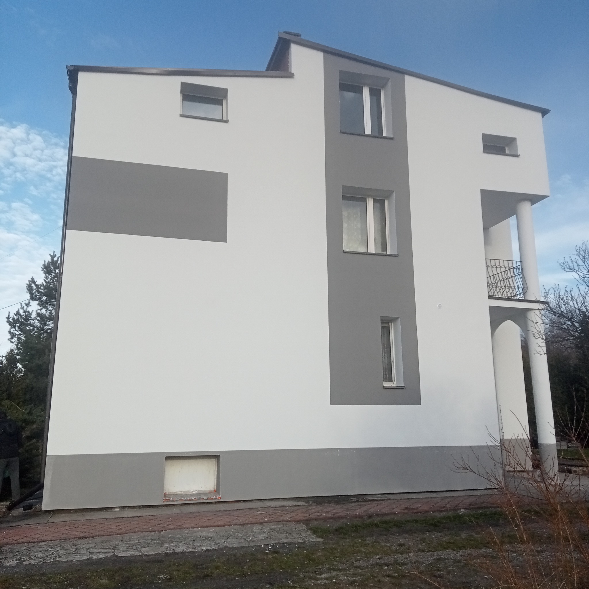 Dwukondygnacyjny dom z elewacją w kolorze białym i szarym, z balkonem z kutą balustradą i kolumnami, widok z zewnątrz, błękitne niebo w tle.