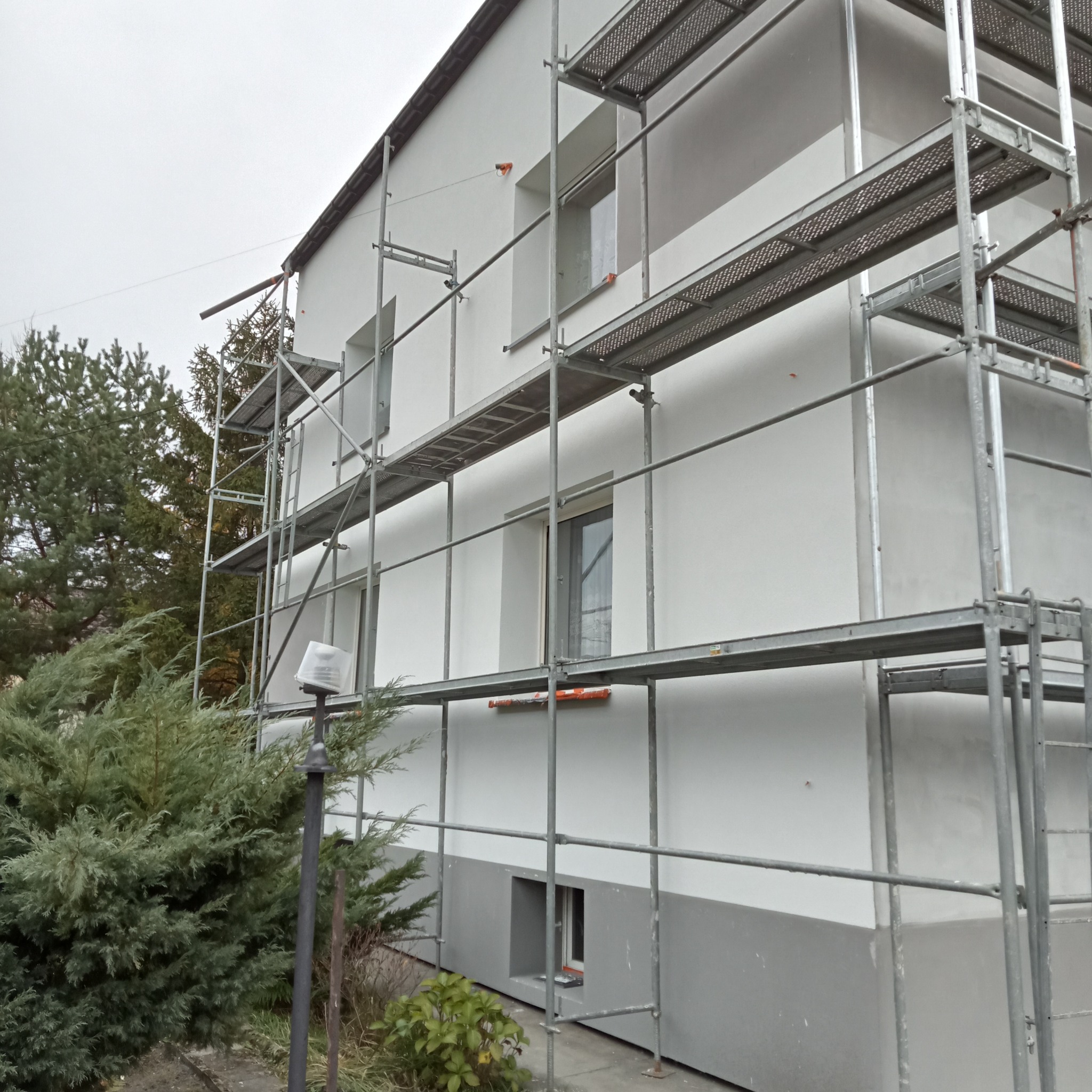 Dwukondygnacyjny budynek w trakcie prac elewacyjnych, otoczony rusztowaniem i zielenią, z widocznym fragmentem pomalowanej na szaro dolnej części elewacji.
