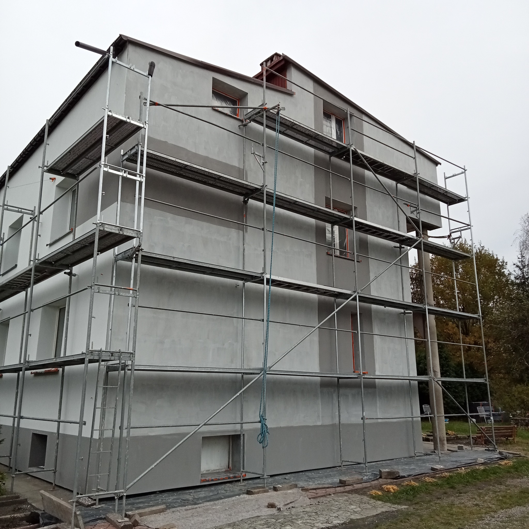 Trzypiętrowy budynek w trakcie prac elewacyjnych, z widocznym rusztowaniem, częściowo otynkowaną i pomalowaną fasadą w odcieniach szarości i bieli, okna zabezpieczone folią.