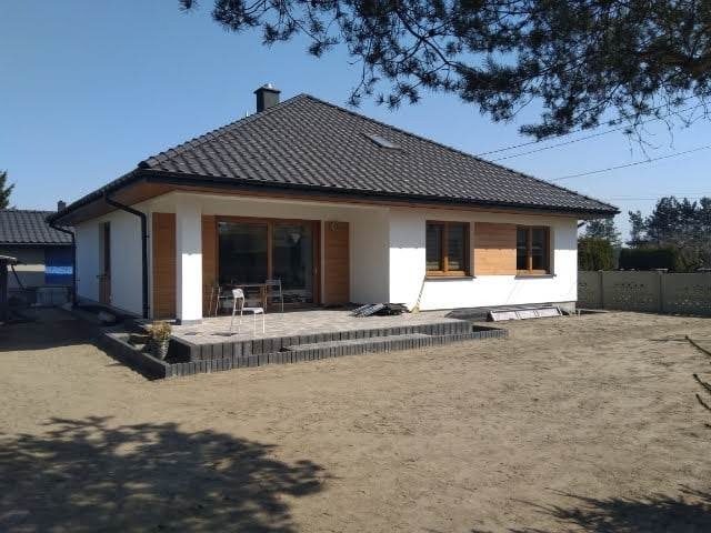 Biały dom jednorodzinny z ciemną dachówką, drewnianymi okiennicami i wykończonym tarasem z kostki, na działce przed domem widoczne ślady prac ziemnych.