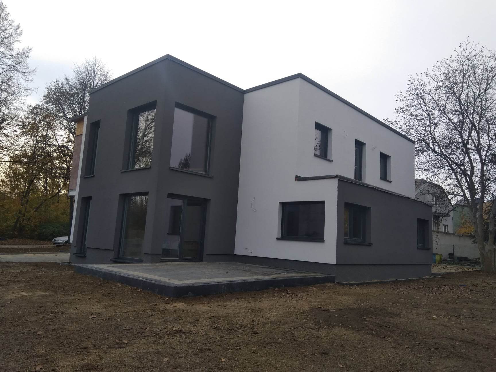 Dwukondygnacyjny dom z elewacją w dwóch odcieniach szarości i bieli, z widocznym tarasem wyłożonym szarą kostką, w jesiennej scenerii.