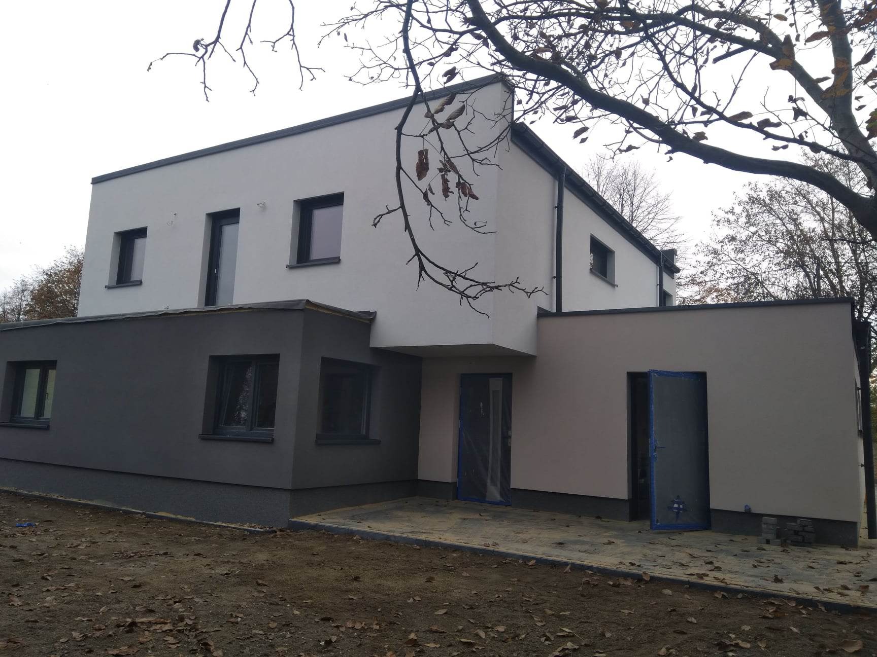 Nowoczesny dom z elewacją w odcieniach szarości i bieli, z widocznymi zabezpieczeniami na drzwiach, sugerującymi świeżo zakończone prace tynkarskie.