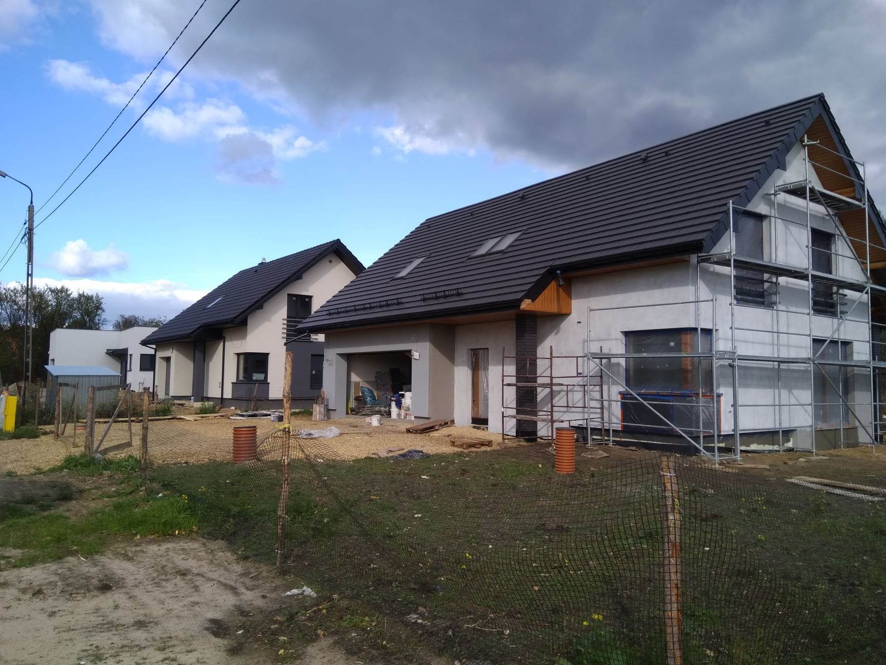 Dwa nowoczesne domy jednorodzinne w trakcie wykończenia elewacji; jeden z domów otoczony rusztowaniem, okna zabezpieczone folią, widoczne worki z materiałami budowlanymi w garażu.
