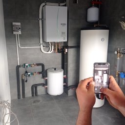 NOVEL ENERGY SP. Z O.O. - Instalacja grzewcza z zasobnikiem CWU, pompą ciepła i rozbudowaną armaturą, fotografowana telefonem komórkowym na tle szarych płytek.
