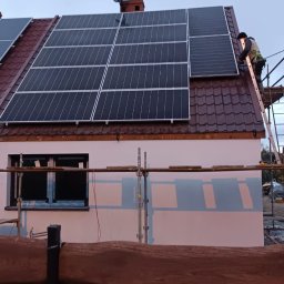 NOVEL ENERGY SP. Z O.O. - Montaż paneli fotowoltaicznych na dachu domu z brązową dachówką, widoczny pracownik na drabinie i rusztowanie przy ścianie. Na dachu antena satelitarna.
