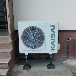 NOVEL ENERGY SP. Z O.O. - Zewnętrzna jednostka klimatyzacji Kaisai zamontowana na metalowym stelażu, ustawiona obok schodów wejściowych i kranu z niebieską rurą.