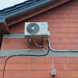 NOVEL ENERGY SP. Z O.O. - Zewnętrzna jednostka klimatyzacji zamontowana na ceglanej ścianie budynku z widocznymi przewodami i elementami mocującymi.