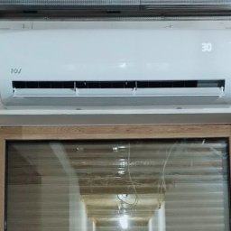 NOVEL ENERGY SP. Z O.O. - Biały klimatyzator ścienny z wyświetlaczem ustawionej temperatury, zamontowany nad oknem z drewnianą ramą i roletami poziomymi.