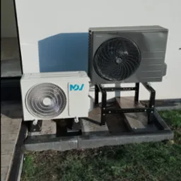 Dwie jednostki zewnętrzne klimatyzacji/wentylacji, jedna biała z niebieskim logo, druga szara Panasonic, zamontowane na metalowych wspornikach na zewnątrz budynku z trawnikiem.