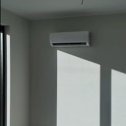 NOVEL ENERGY SP. Z O.O. - Biała jednostka klimatyzacji zamontowana na jasnej ścianie w nowym pomieszczeniu, widoczne puszki elektryczne i resztki opakowań na podłodze, oświetlona silnym światłem słonecznym wpadającym przez...