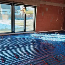 Niebieskie rury ogrzewania podłogowego ułożone na folii w surowym wnętrzu z ceglanymi ścianami i dużymi oknami z widokiem na zaśnieżony ogród.