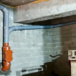 NOVEL ENERGY SP. Z O.O. - Szare rury kanalizacyjne z pomarańczowym zaworem zwrotnym zamocowane do ściany z cegieł w piwnicy. Widoczne mocowania i fragment palety w rogu.