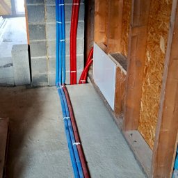 NOVEL ENERGY SP. Z O.O. - Ułożone, niebieskie i czerwone rury instalacyjne, przymocowane do ściany z bloczków i drewnianej konstrukcji. Widoczny fragment podłogi i biała skrzynka instalacyjna.