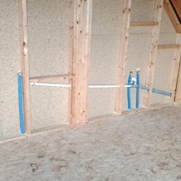 NOVEL ENERGY SP. Z O.O. - Surowy montaż instalacji wodnej w drewnianej konstrukcji szkieletowej poddasza. Niebieskie rury i białe przewody, widoczne przyłącza. Etap prac przed wykończeniem.