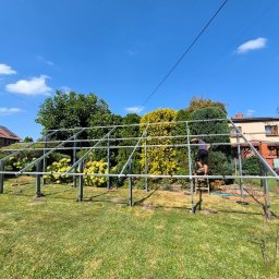 NOVEL ENERGY SP. Z O.O. - Montaż konstrukcji wsporczej pod panele fotowoltaiczne w ogrodzie. Widoczny monter na drabinie, metalowa konstrukcja na tle zieleni i domu z panelami na dachu.