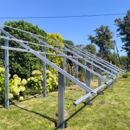 NOVEL ENERGY SP. Z O.O. - Montaż stelażu pod panele fotowoltaiczne w ogrodzie, widok z dołu. Metalowa konstrukcja na trawniku, pracownik na drabinie. Błękitne niebo w tle.