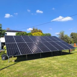NOVEL ENERGY SP. Z O.O. - Panel fotowoltaiczny na trawniku, odbijający słońce. Instalacja na metalowych nogach, dom w tle, błękitne niebo. Widok z poziomu trawy, słoneczny dzień.