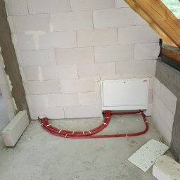 NOVEL ENERGY SP. Z O.O. - Instalacja rur w budynku w trakcie budowy. Czerwone rury prowadzone wzdłuż ściany z białych bloków, pod białą skrzynką. Widoczne drewniane elementy konstrukcji dachu.