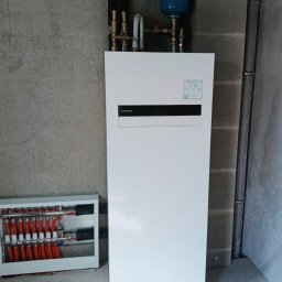 NOVEL ENERGY SP. Z O.O. - Biała pompa ciepła Panasonic Aquarea Smart Cloud z niebieskim zbiornikiem wyrównawczym i rozdzielaczem ogrzewania podłogowego na tle surowej ściany.