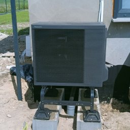 NOVEL ENERGY SP. Z O.O. - Zewnętrzna jednostka pompy ciepła Panasonic Aquarea A, zamontowana na betonowych bloczkach przy ścianie budynku w trakcie instalacji. Widoczne przewody i elementy montażowe.