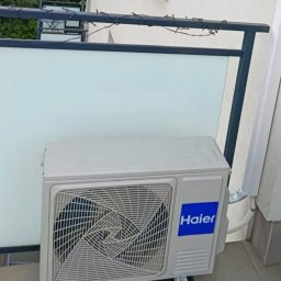 NOVEL ENERGY SP. Z O.O. - Klimatyzator Haier na balkonie, zamontowany na czarnych podstawkach antywibracyjnych. Widoczne rury odprowadzające. Balkon z białą balustradą i czarną ramą.