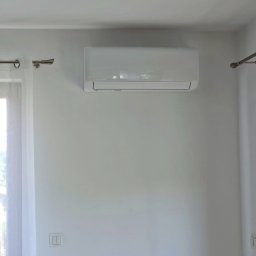NOVEL ENERGY SP. Z O.O. - Biały klimatyzator AUX zamontowany na białej ścianie w jasnym pomieszczeniu. Widoczny fragment okna z firanką i włącznik światła.