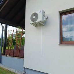 NOVEL ENERGY SP. Z O.O. - Biała jednostka zewnętrzna klimatyzacji AUX zamontowana na ścianie domu z widoczną instalacją. Obok okno z brązową ramą i fragment zadaszonego tarasu.