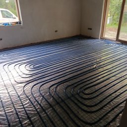 NOVEL ENERGY SP. Z O.O. - Niebieskie rury ogrzewania podłogowego ułożone na folii w surowym wnętrzu z oknami. Widoczne złącza i mocowania rur. Etap instalacji przed wylewką.