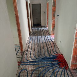 NOVEL ENERGY SP. Z O.O. - Korytarz z ułożonym systemem ogrzewania podłogowego: niebieskie rury na siatce, czerwone złączki przy ścianie. Surowy stan wykończenia ścian i futryn.