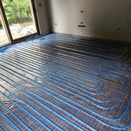NOVEL ENERGY SP. Z O.O. - Niebieskie rury ogrzewania podłogowego ułożone na folii z czerwoną siatką w surowym wnętrzu. Widok z góry na instalację przed wylaniem posadzki.