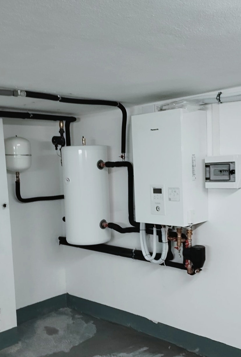 Biała pompa ciepła Panasonic z zasobnikiem ciepłej wody, czarnymi rurami izolowanymi i rozdzielnicą elektryczną na ścianie w kotłowni z szarą podłogą.