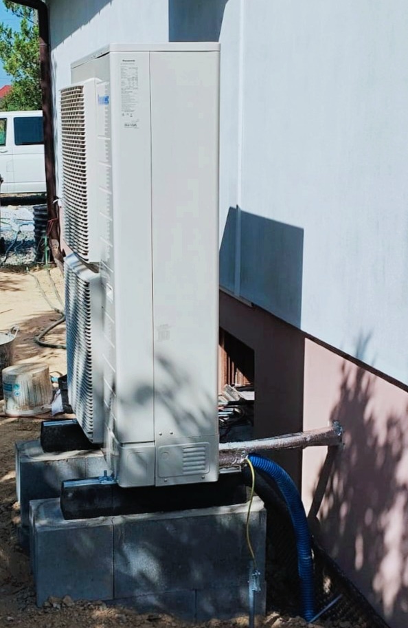 Dwie jednostki zewnętrzne pompy ciepła marki Panasonic, zamontowane na betonowych bloczkach przy ścianie budynku, z widocznymi niebieskimi rurami odprowadzającymi skropliny.