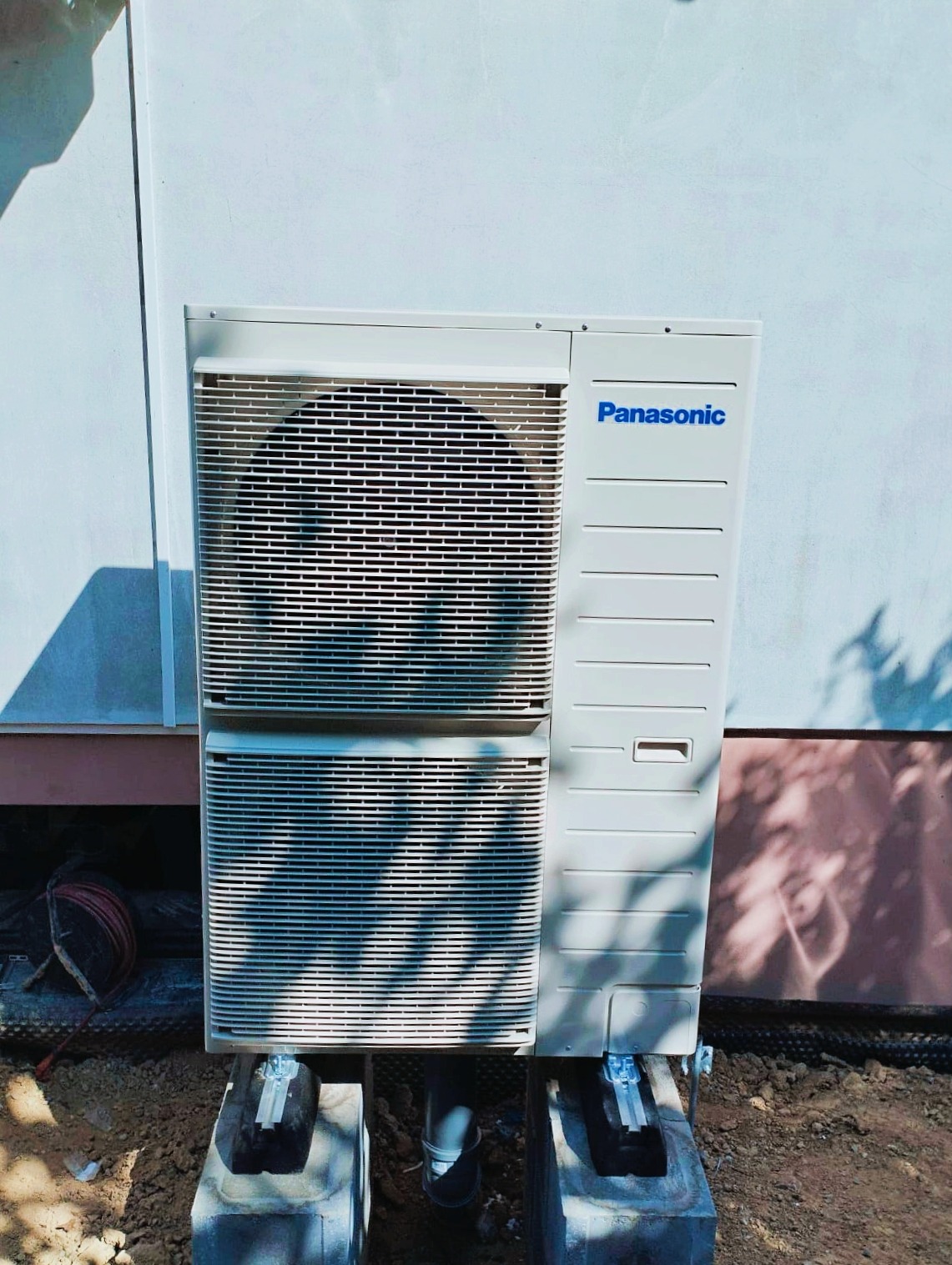 Zewnętrzna jednostka pompy ciepła Panasonic, zamontowana na betonowych bloczkach przy jasnej ścianie budynku, widoczne przewody i fragment instalacji.