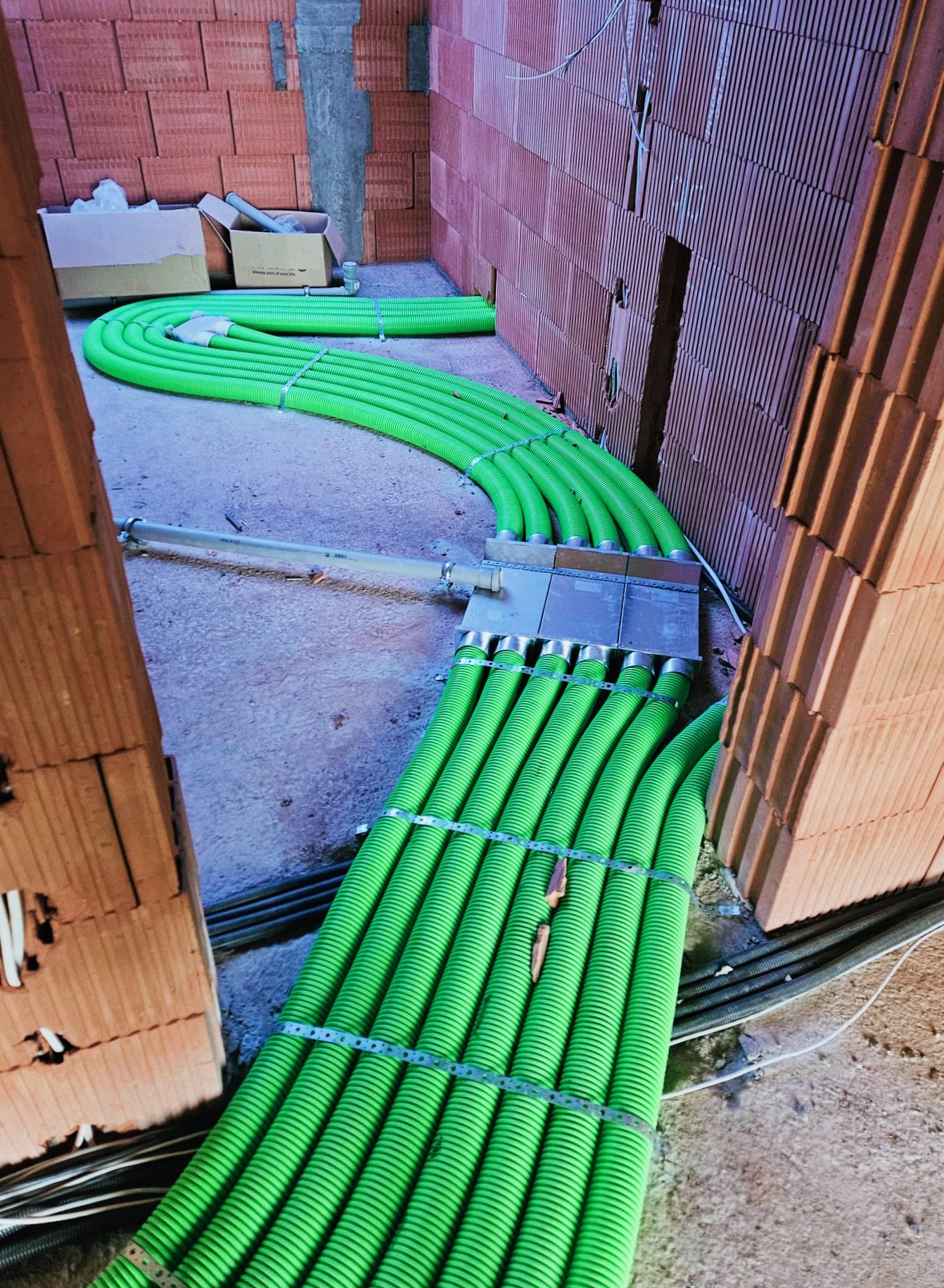 Wnętrze w trakcie budowy z widocznymi zielonymi rurami wentylacyjnymi poprowadzonymi wzdłuż ściany z cegły ceramicznej, zabezpieczonymi metalowymi obejmami. Na podłodze widoczne dodatkowe przewody...