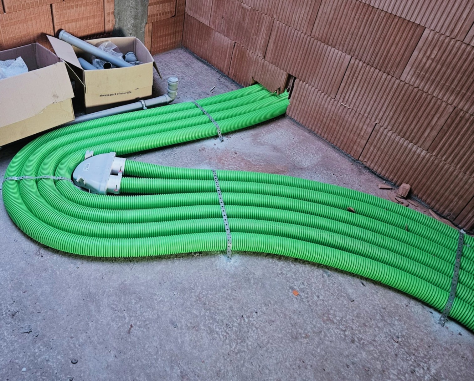 Zielone, karbowane rury prowadzone w podłodze w nowym budynku, przymocowane metalowymi obejmami, z widocznym rozdzielaczem i elementami instalacyjnymi w kartonach w tle.