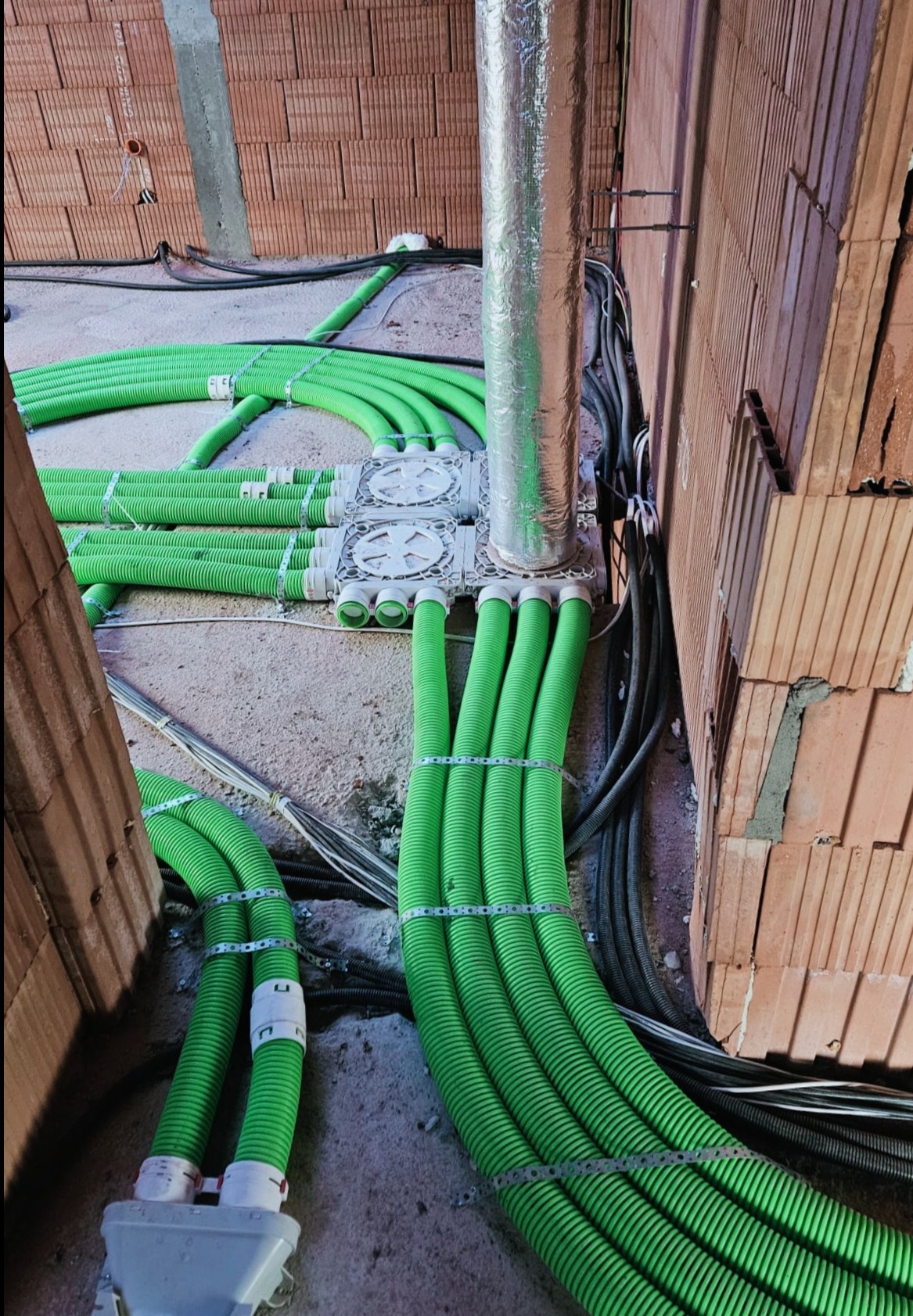 Wnętrze budynku w trakcie budowy z widocznymi zielonymi rurami wentylacyjnymi połączonymi z szarą skrzynką rozdzielczą, oraz srebrną izolowaną rurą pionową, przymocowanymi do ceglanej ściany.