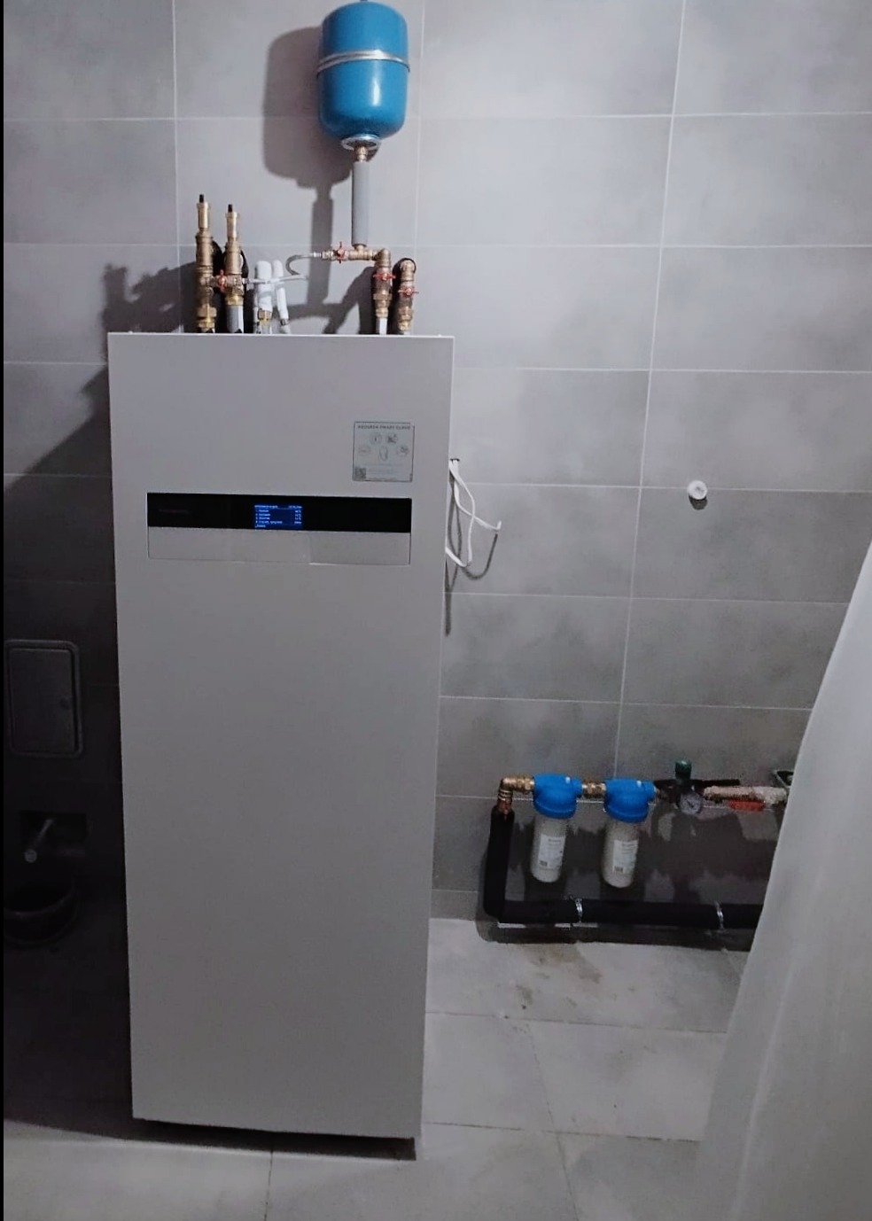 Biała pompa ciepła z niebieskim zbiornikiem wyrównawczym zamontowana na tle szarych płytek, widoczne rury i filtry.