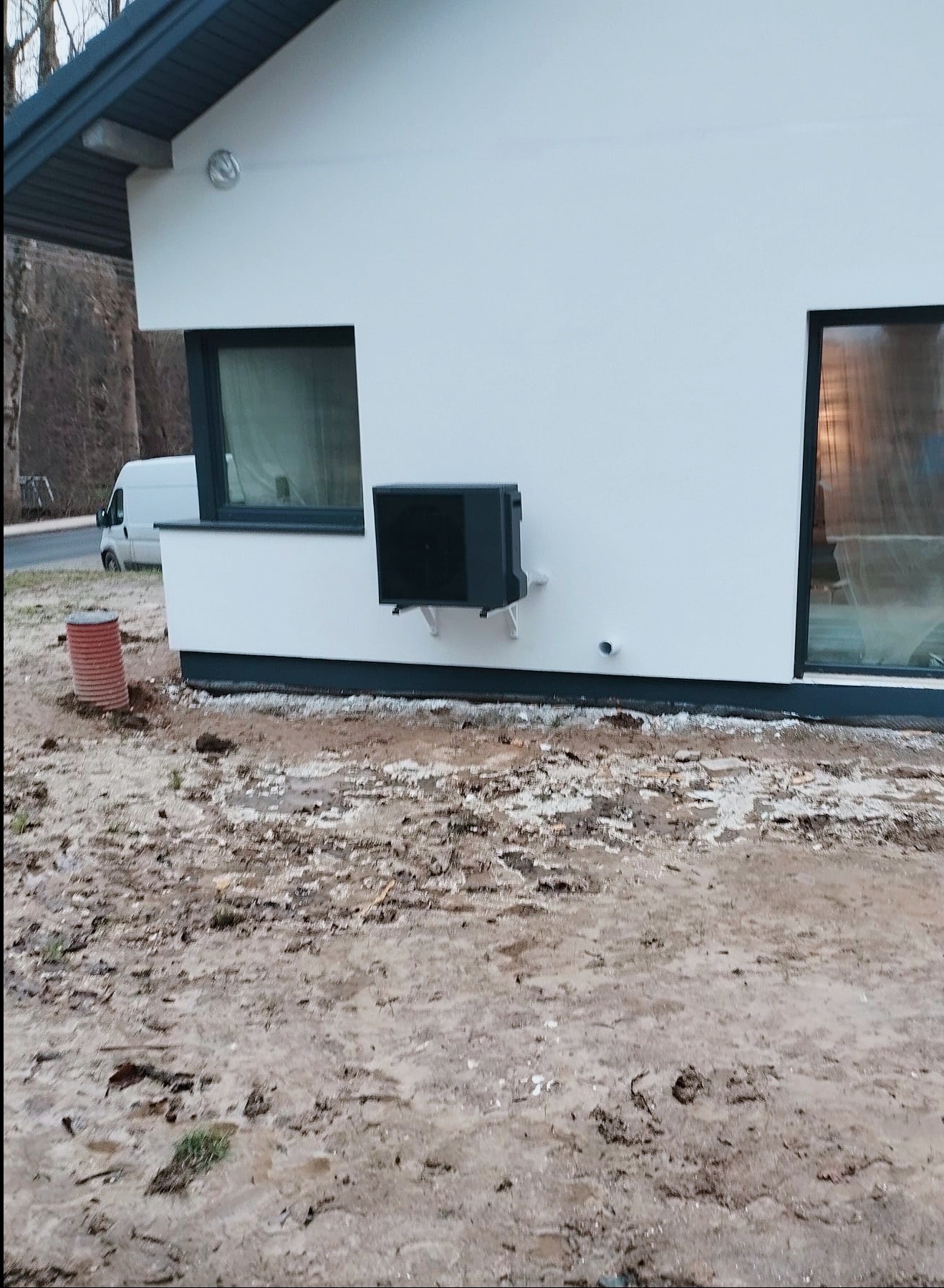 Zewnętrzna jednostka pompy ciepła zamontowana na ścianie nowoczesnego domu z widoczną instalacją i elementami otoczenia w trakcie montażu.